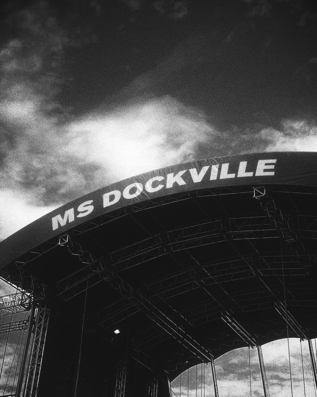 Schwarzweißfotografie des MS Dockville Festivalbühnenbogens von unten — Stahlkonstruktion mit dem Schriftzug "MS Dockville" vor dramatischem Wolkenhimmel — Festivalaufbau, MS Dockville Hamburg
